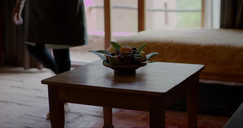 Interior design of a small table with artificial decoration next to a bed and a person in the background picking up an object and leaving the room