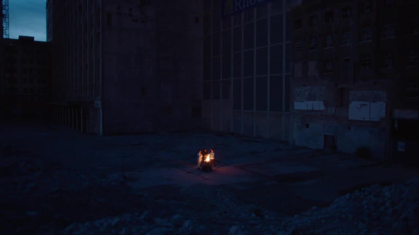 A small group of people huddle around a fire in a dark abandoned industrial space