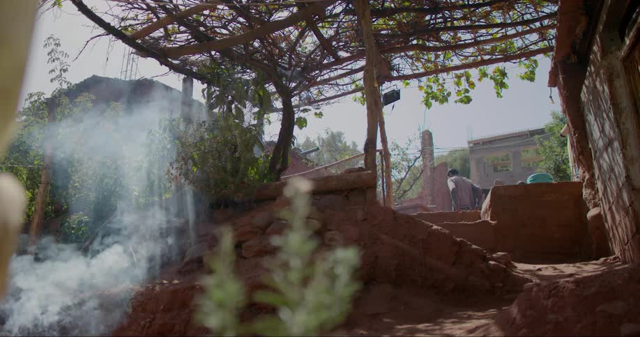 A woman in a red dress walks up a dirt path under a vinecovered pergola with smoke rising nearby