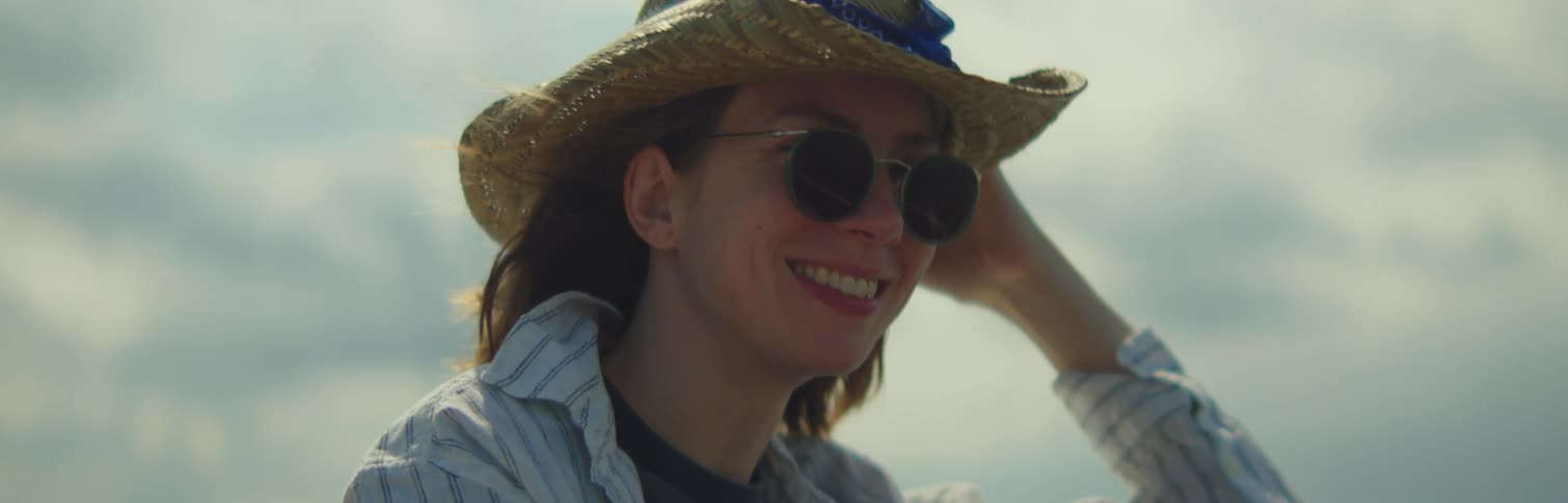 A woman wearing a hat sunglasses and a shirt looks around and smiles against the cloudy sky