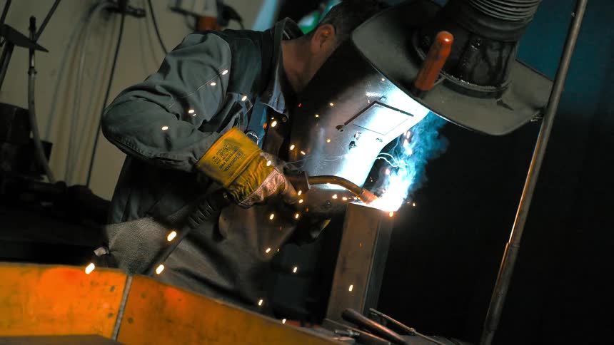 A welder in a dark workshop uses a welding torch to join two pieces of metal