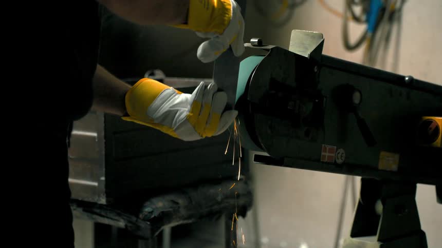 Closeup of a persons hands wearing yellow gloves using a bench grinder to sharpen a piece of metal