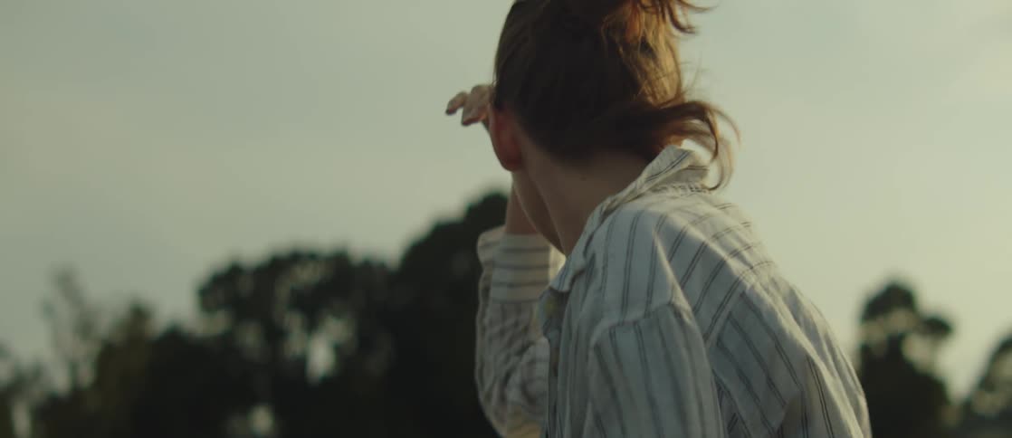 Woman with hand on forehead pointing in a direction while standing in an outdoor setting with trees in the background