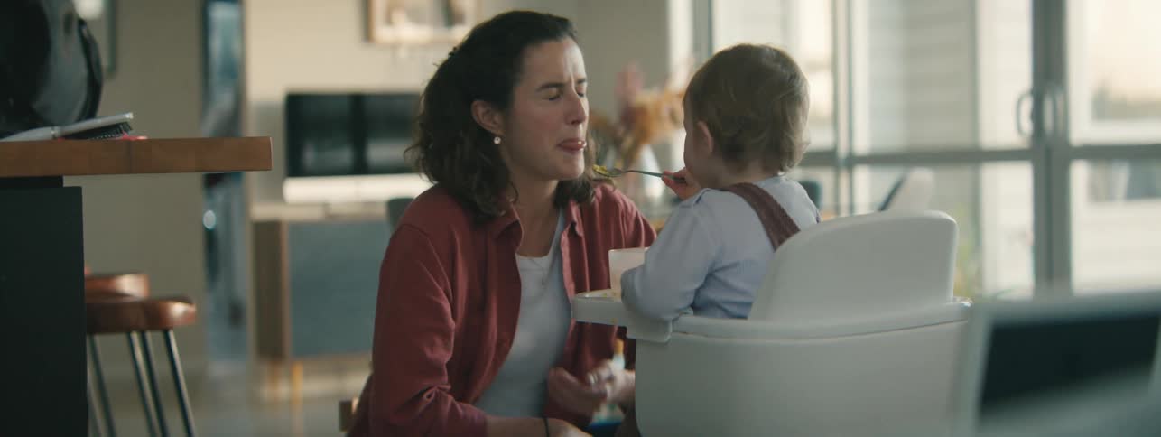 A mother crouches next to a child seated in a high chair playing with the child and his food