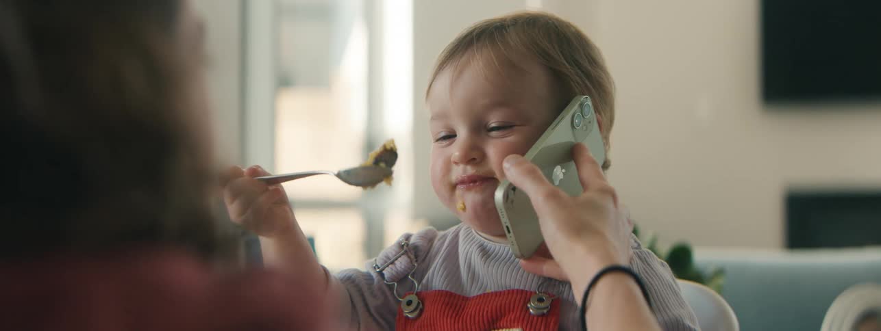 A mother holding a phone to her toddler daughters ear while being fed with a spoon creating a playful and wholesome moment