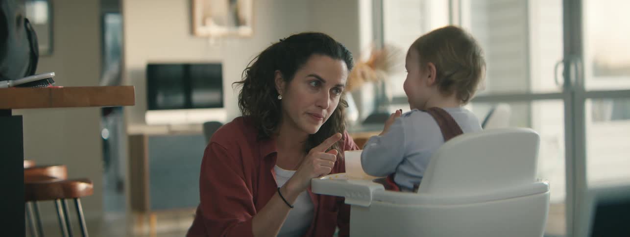 A mother crouches next to a child seated in a high chair playing with the child and his food