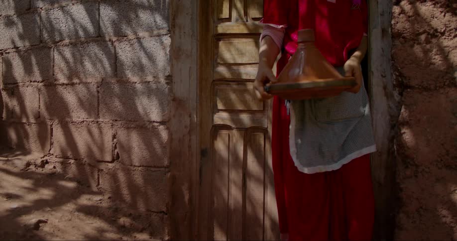 Person in a red apron coming out of a simple house carrying a plate in a bowl