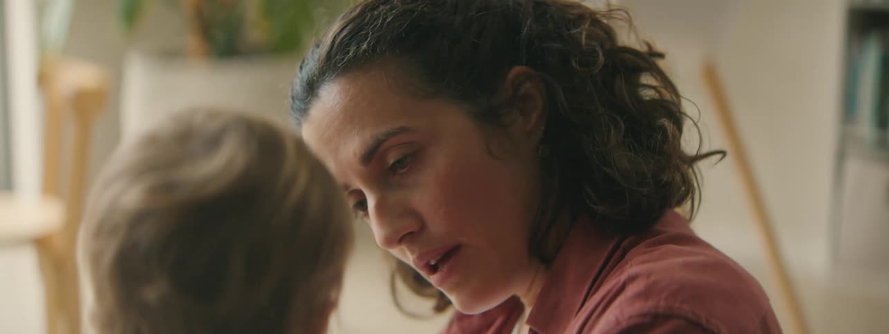 A mother with curly hair and a red shirt gently caresses her toddler daughter