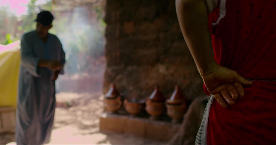 Two people wearing robes outdoors with several clay pots lined up against a rustic wall