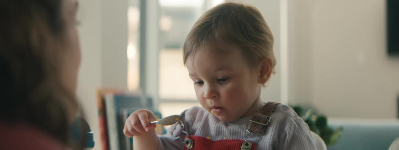 A mother watches her toddler son eat cereal in a kitchen