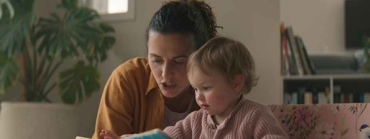 A mother shows her toddler son a book while theyre both sitting on a couch in a living room