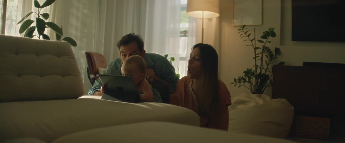 A family with a toddler sits together in the living room gathered around a tablet