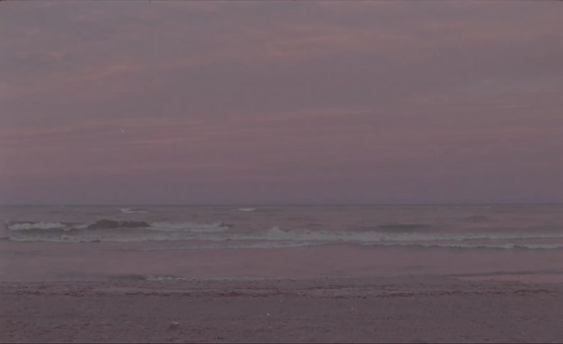A 16mm vast ocean stretches out under a hazy pink sky while gentle waves lap at the shore of a sandy beach