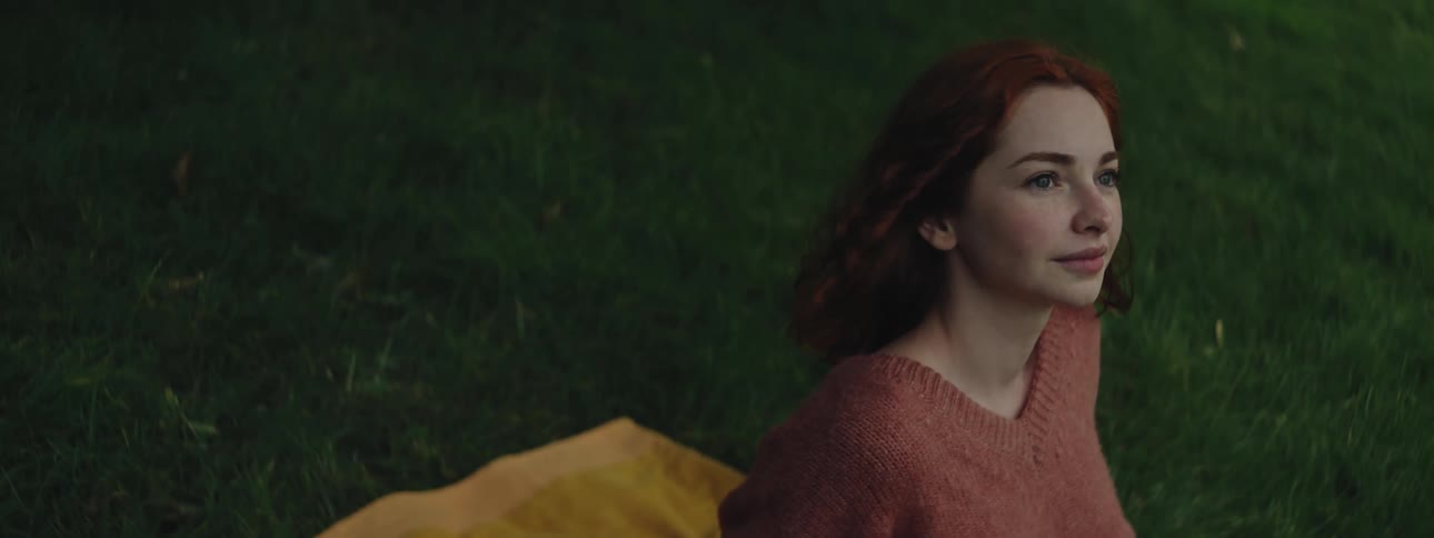 A red haired teenage girl wearing a sweater smiles while sitting on a yellow towel outdoors