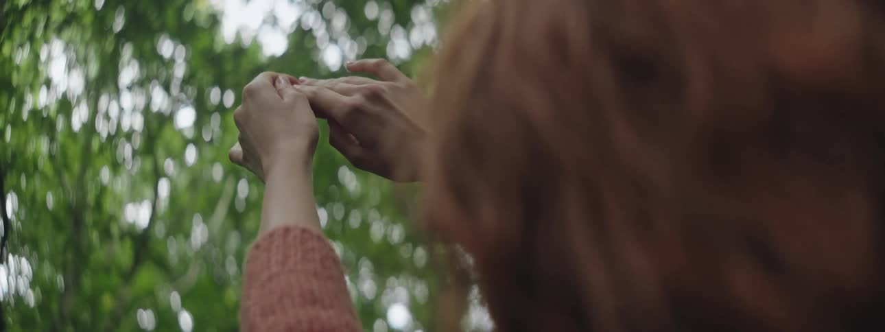 Red haired woman with her hands pointing up against a background of green leaves of a tree