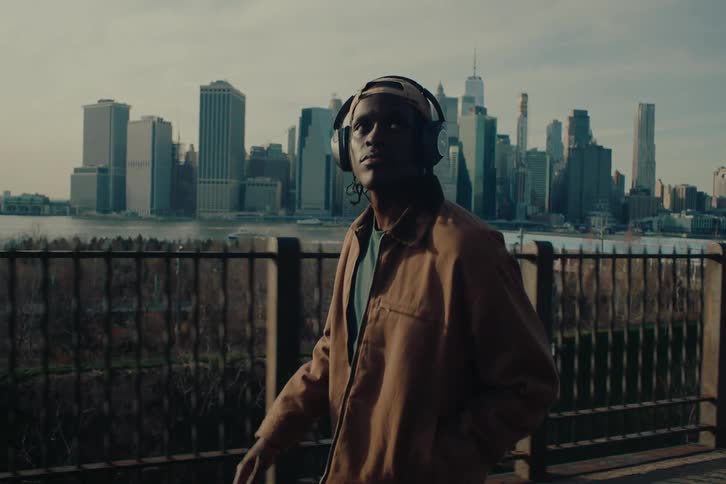 A young man wearing a brown jacket and headphones walks in a park on the outskirts of new york city