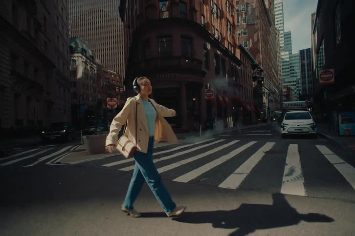 A woman in a light beige jacket crosses a city street wearing headphones and listening to music