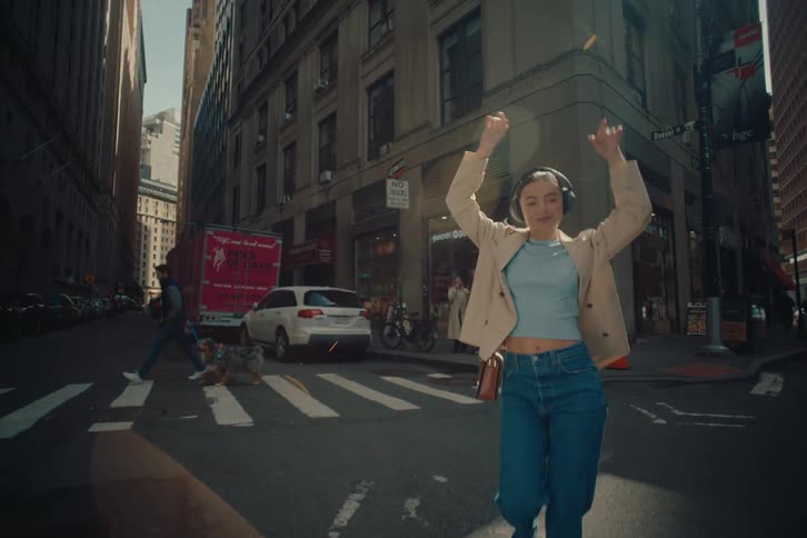 A woman in a light beige jacket crosses a city street wearing headphones and listening to music