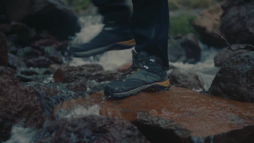 Person wearing a pair of boots walks across a wet rock in the middle of a small river