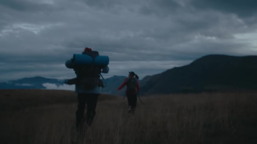 Two hikers running through an open field at dusk with large backpacks and rolled sleeping mats strapped to them