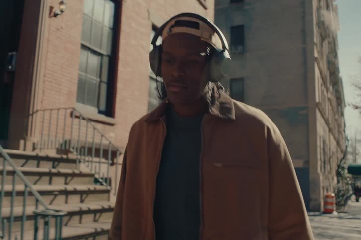 A young man wearing a brown coat listens to music on large headphones while walking in the city
