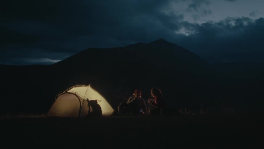 Silhouette of two people sitting next to a tent and a mountain in the background in the dark of night