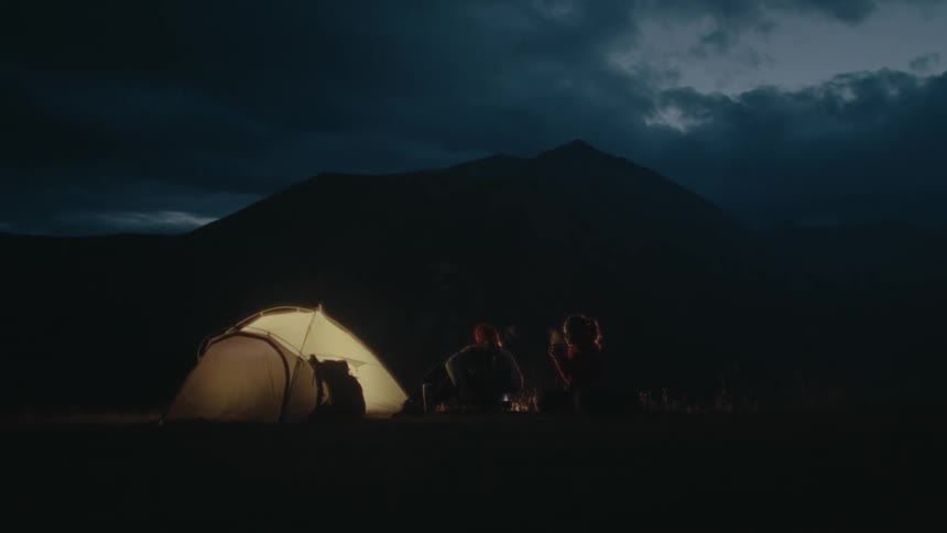 Silhouette of two people sitting next to a tent and a mountain in the background