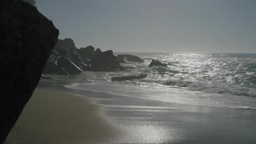 Beach with gentle waves crashing on the sand and large rocks in the background under a clear sky