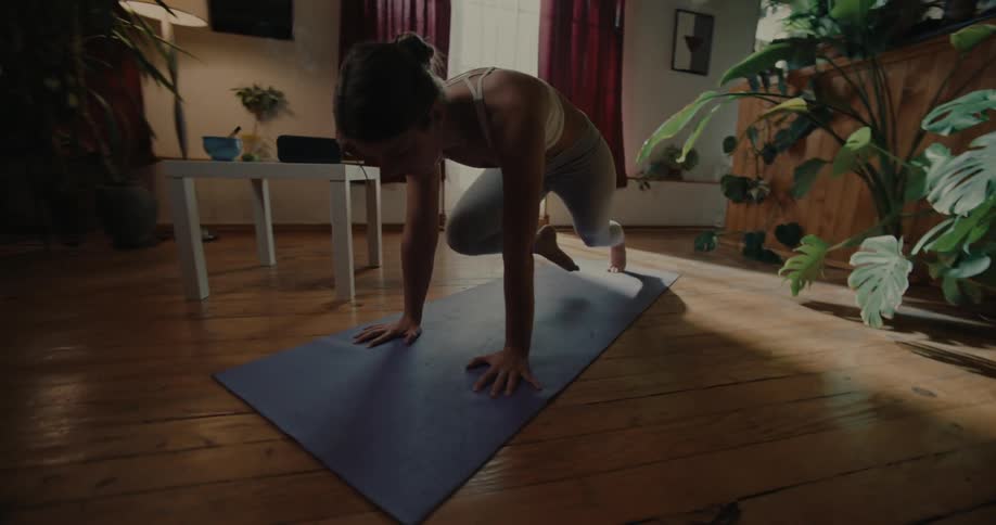 A woman in a sports bra and gray leggings performs a mountain climber exercise in a room filled with houseplants