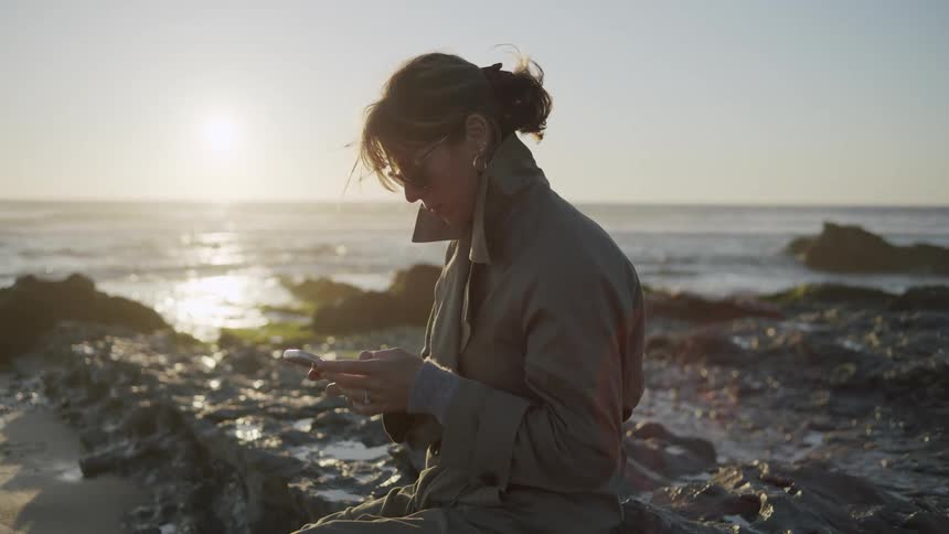 Woman with sunglasses sitting on rocks by the sea interacting with a smartphone