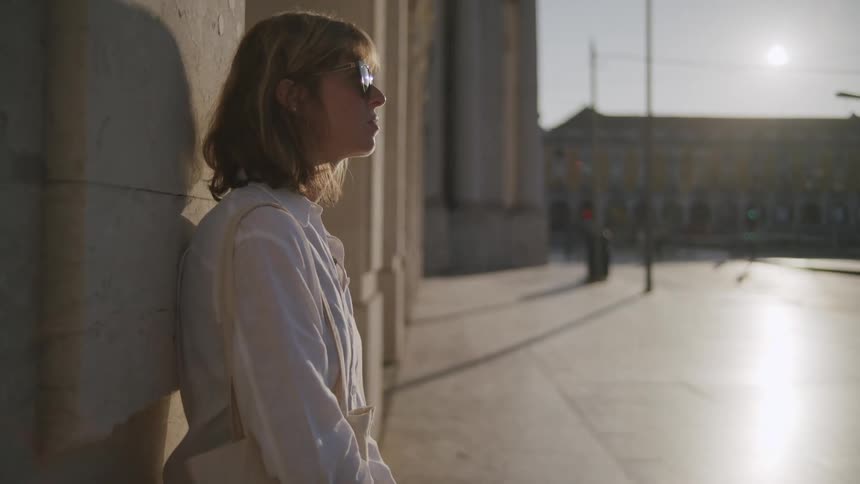 Woman in a white shirt and sunglasses leaning against a wall in a public place