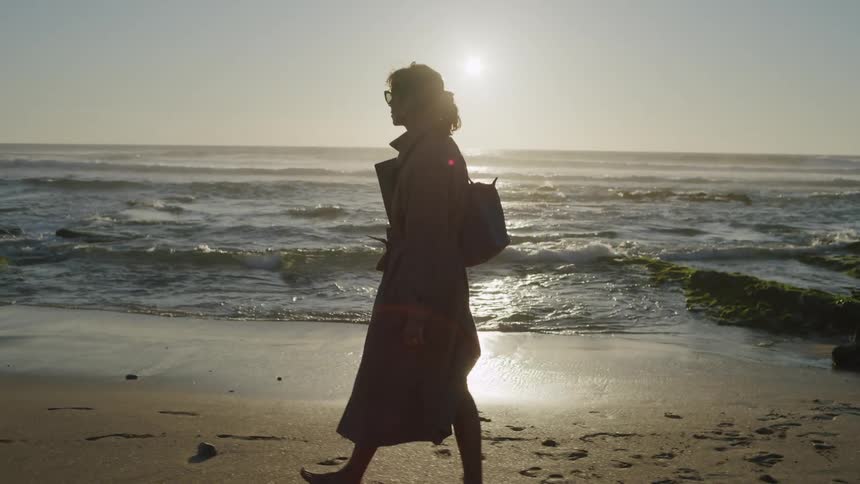 A woman in a trench coat walks on a beach as the sun hangs low in the sky