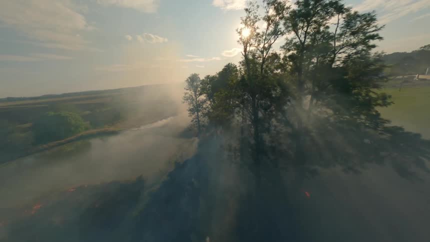 An aerial view of a field fire with smoke rising into the air and sunlight behind trees