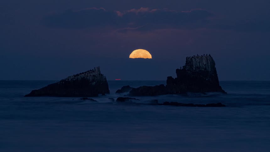 A full moon sinks below the horizon as waves move around a rock formation in the ocean