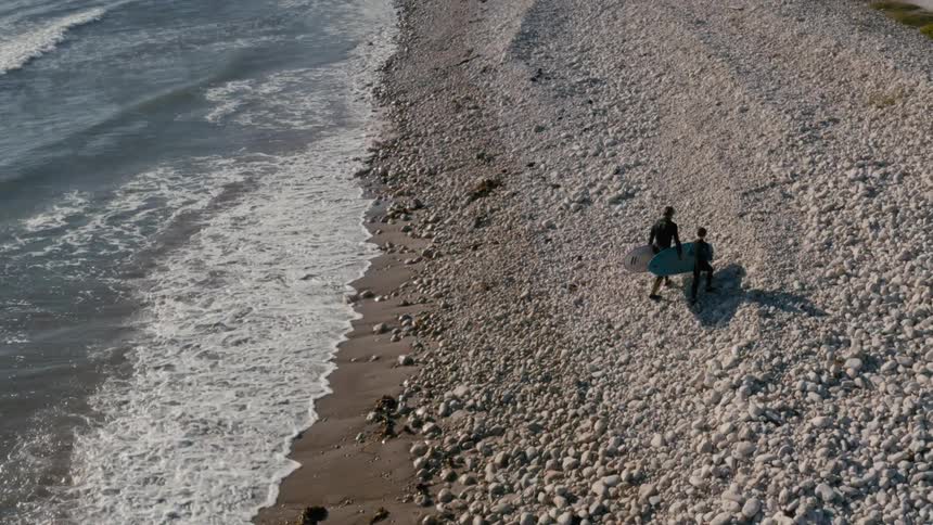An aerial view of two surfers carrying surfboards walking away from the coast on a pebblefilled shoreline