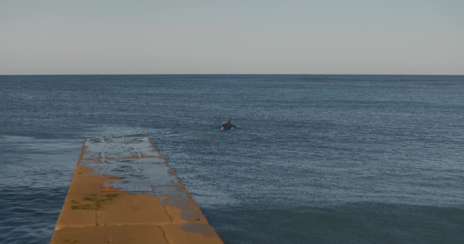 A lone surfer lays on their board paddling away from a concrete jetty