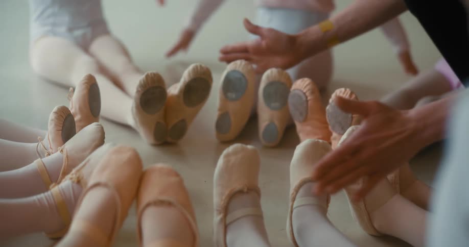 Closeup of the feet of several ballerinas sitting together and an adults hand helping