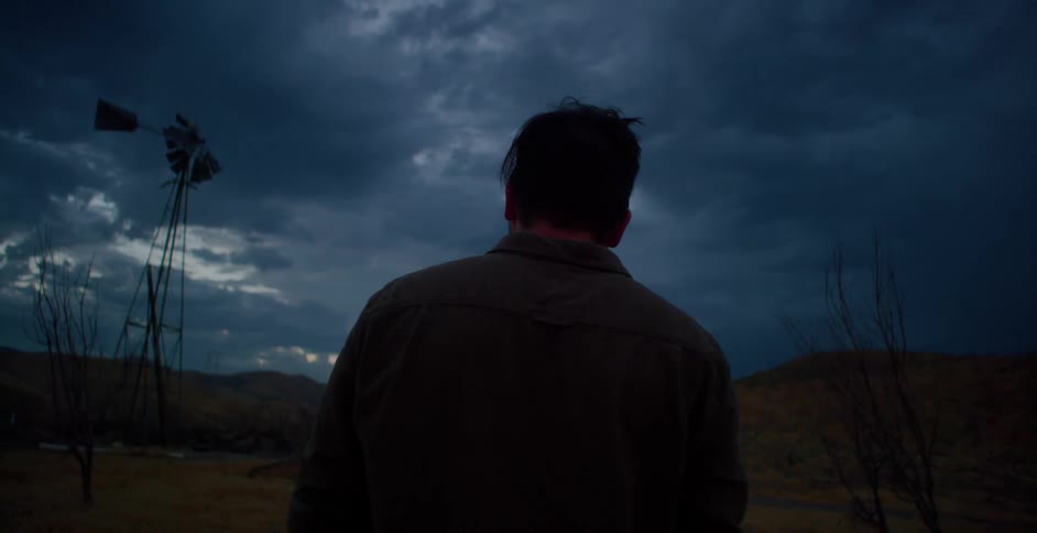 A man wearing a cowboy hat takes it off and looks at a windpump under a cloudy sky at dusk