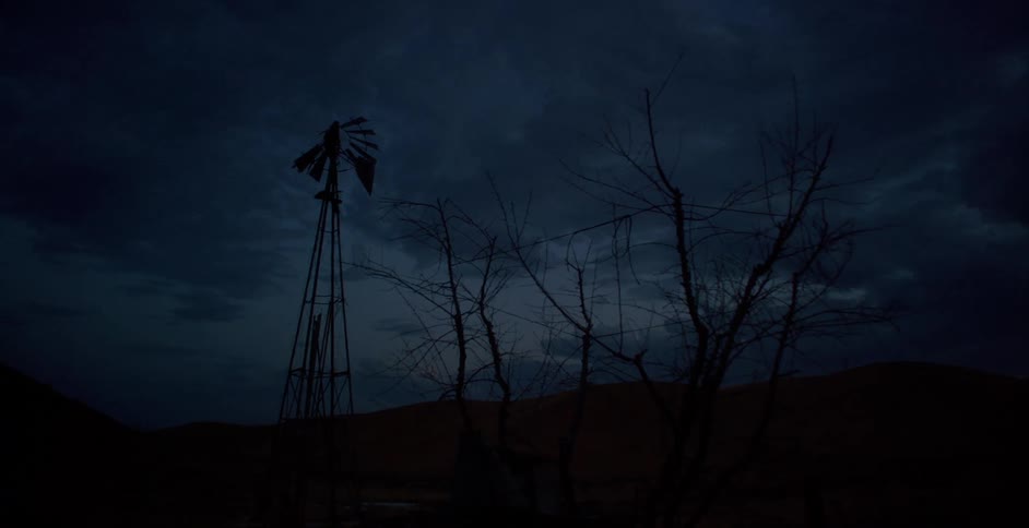 A broken windmill and dead trees on a farm at night