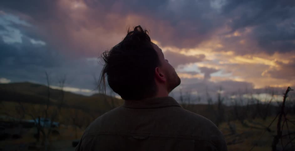 Back view of a man taking off his hat and looking at the cloudy dawn sky while standing in a field