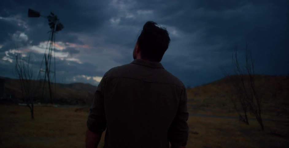 A cowboy stands outside on a farm and looks at storm clouds over mountains