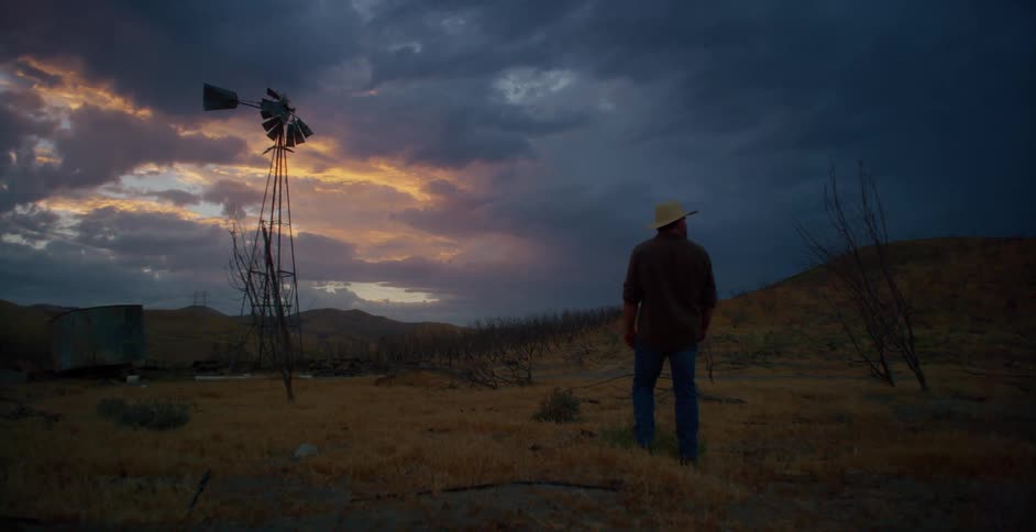 A solitary rancher in a cowboy hat walks on the ranch under a cloudy sky at dusk