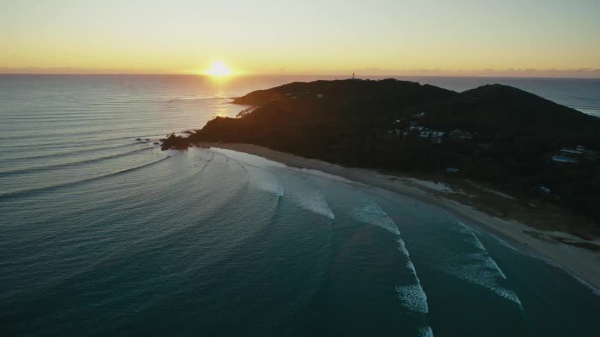 Aerial view of the sun setting over a coastal landscape casting long shadows over the beach and the ocean waves