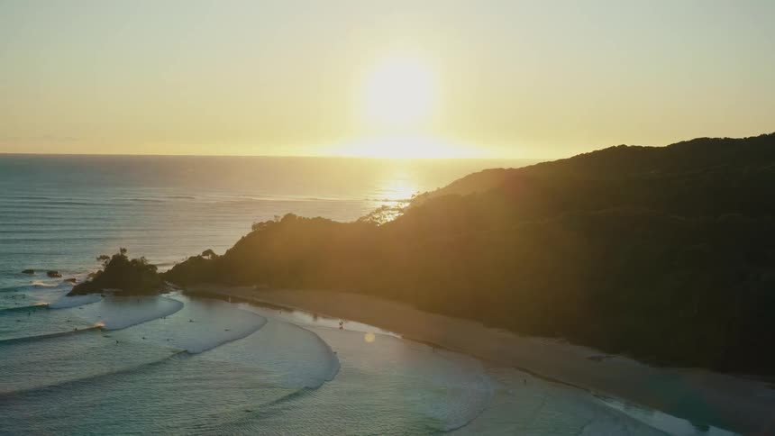 Bright sun sitting on the horizon shines across the ocean onto a forested mountain overlooking the beach