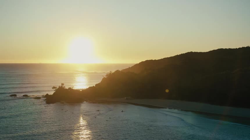 Aerial view of the sun setting over a coastal landscape casting long shadows over the beach and the ocean waves