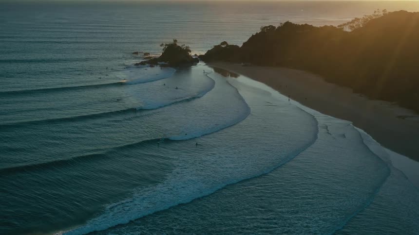 The sun casts a golden glow on the horizon as surfers ride the waves at a secluded beach surrounded by lush green hills and rocky cliffs