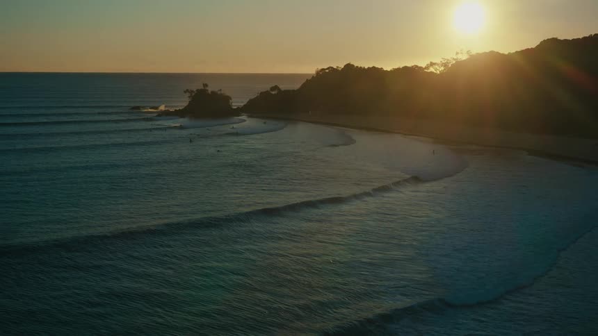 Aerial view of the sun setting over a coastal landscape casting long shadows over the beach and the ocean waves