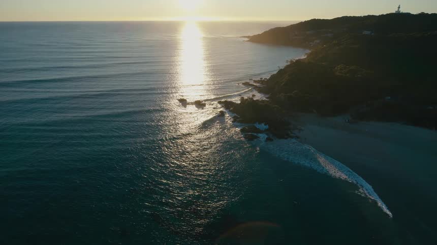 G aerial view of a coastline bathed in the golden light of a setting sun with the ocean sparkling and the rocky shore contrasting against the soft white sand of a small beach
