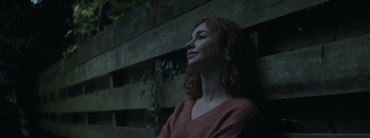 A teenager with red hair leans against a wooden fence outdoors