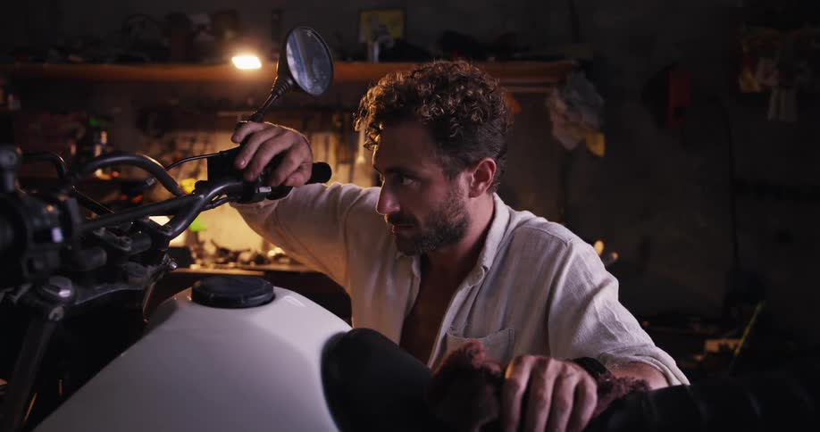 A man in a dimly lit workshop intently examines a motorcycle adjusting its handlebar with focused precision surrounded by tools and mechanical components